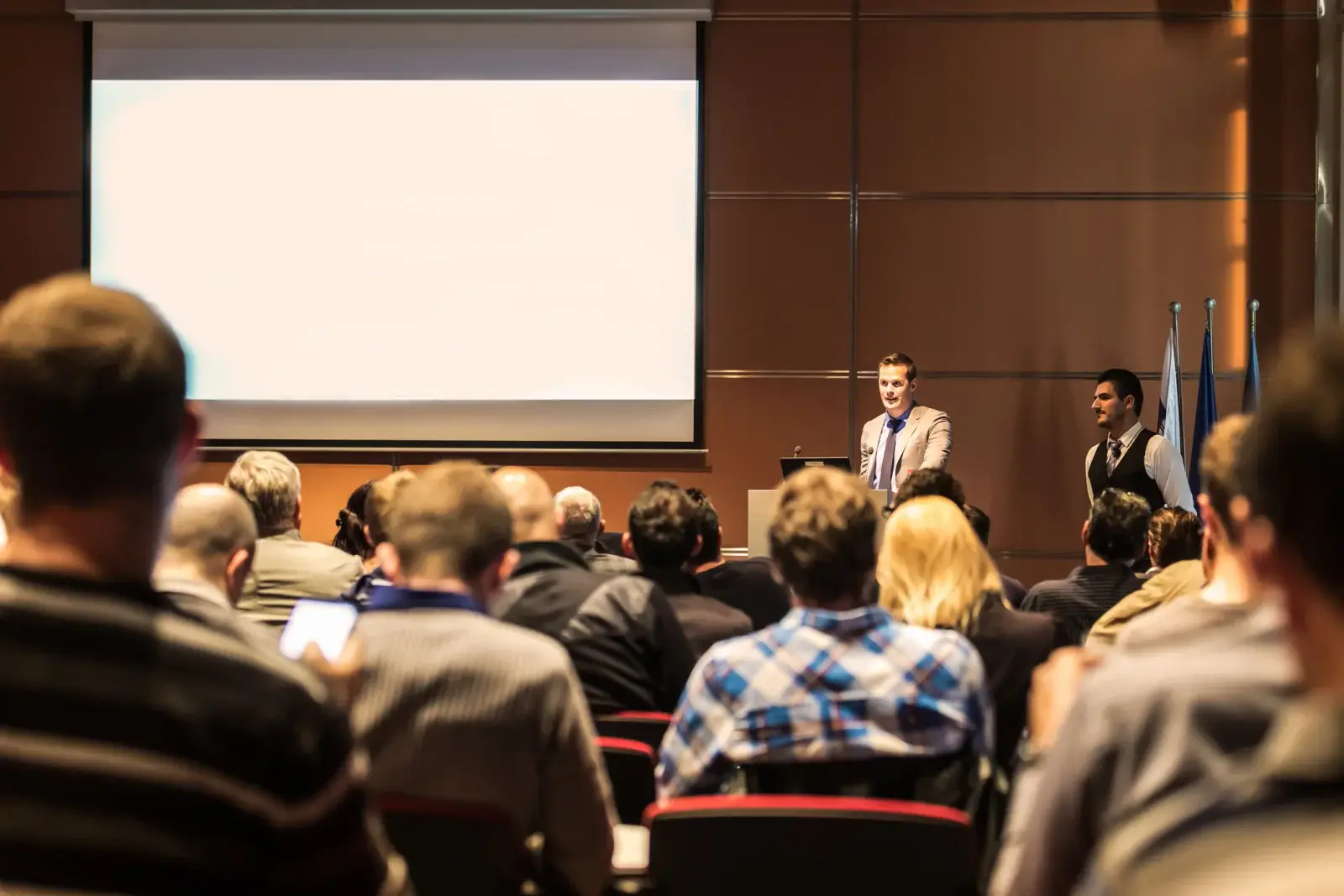 Audience watching presenters beside a large front projection screen