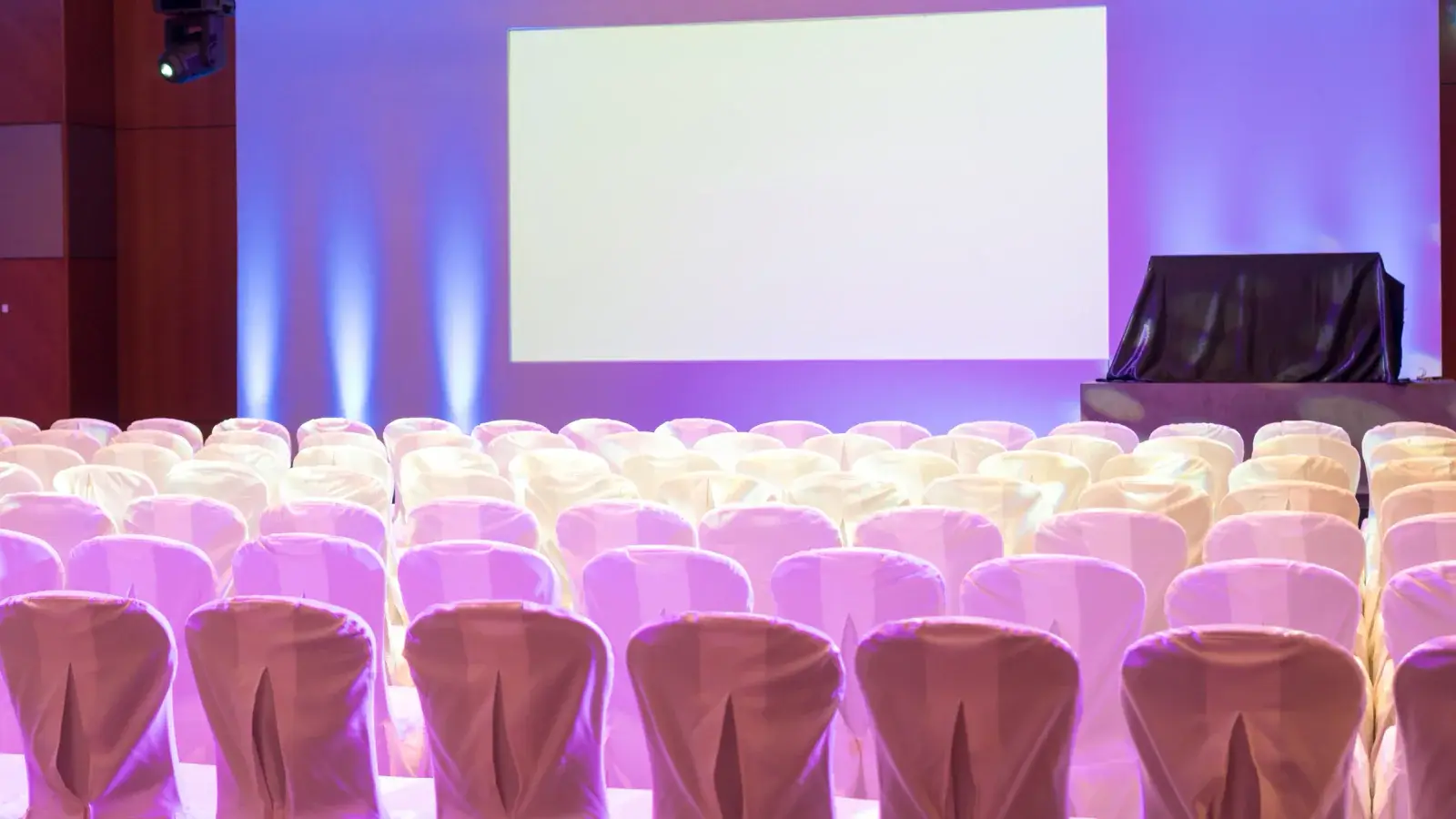 Ballroom with rows of covered chairs facing a large projection screen under purple uplighting